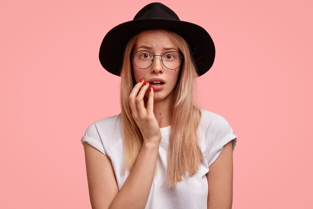 Worried beautiful young woman holds hand near mouth, looks with embarrassed expression, weas hat and t shirt