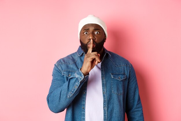 Worried Black man asking to keep quiet, sharing secret and hushing at you, holding finger pressed to lips and staring nervously at camera, pink background