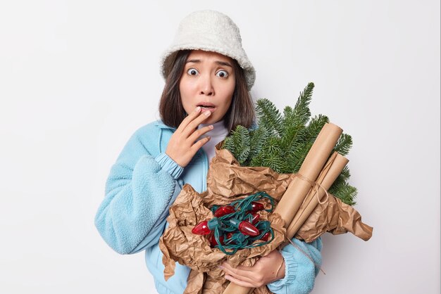 Worried brunette Asian woman trembles and looks embarrassed at camrera forgot to buy something important for winter holidays poses with New Year attributes wears white panana and blue jacket