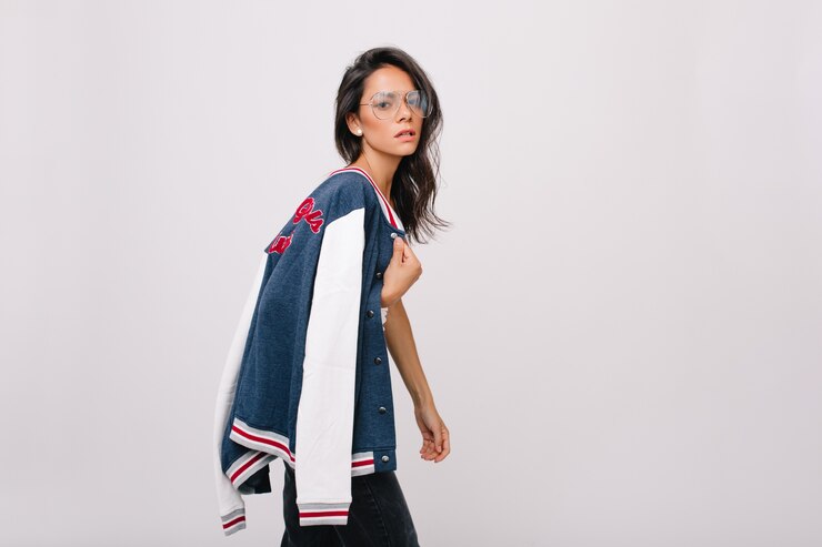 無料Photo カラフルなスポーツジャケットを肩に運ぶ大きなメガネで心配しているブルネットの若い女性。疲れた表情でポーズをとる光沢のある髪の不幸なヨーロッパの女の子。