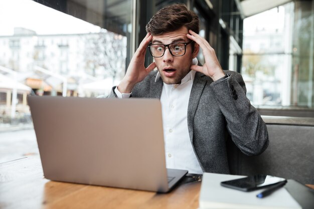 Worried confused businessman in eyeglasses sitting by the table in cafe while holding head and looking at laptop computer