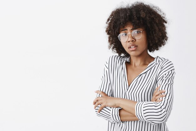 worried dark-skinned female model with curly hair in glasses and office striped blouse holding hands on chest frowning form empathy and anxiety over gray wall