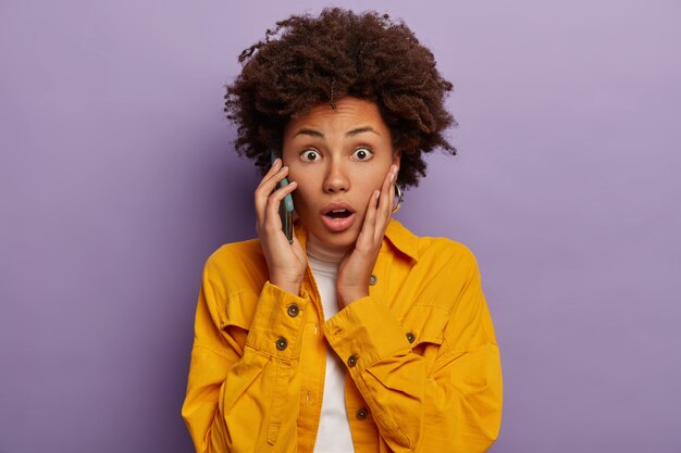 Обеспокоенная темнокожая женщина в панике разговаривает по сотовому телефону, узнает плохие новости, носит желтую рубашку, держит смартфон возле уха
