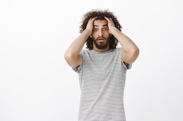 Worried desperate aduly male freelancer with afro haircut in trendy striped t-shirt, holding hands on head and lifting eyebrown, feeling nervous and shocked