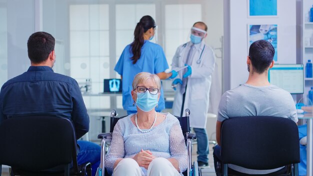 Worried disabled senior woman sitting in wheelchair in hospital waiting area for doctor examination. Old woman wearing face mask against coronavirus infection.