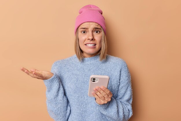 Worried displeased young European woman raises palm looks displeased wears pink hat casual blue jumper holds mobile phone chats online isolated over beige background feels upset after quarrel