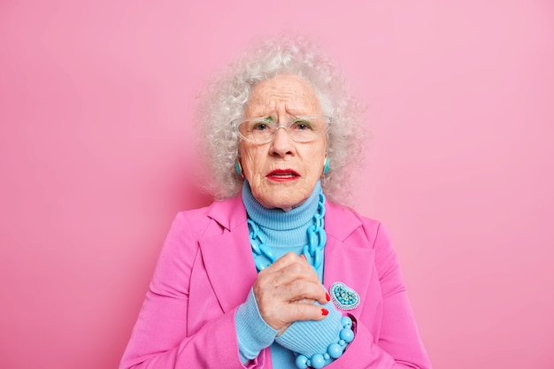 Free photo worried elderly woman with wrinkled face keeps hands together looks disappointed feels nervous about something wears spectacles fashionable clothes