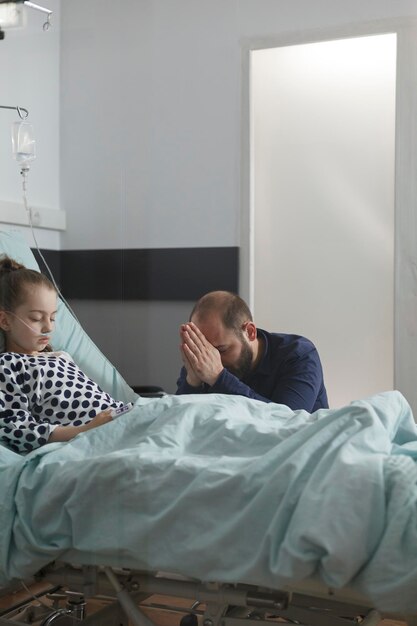 Worried father praying for sick daughter resting on patient bed. Ill little girl under medicine treatment resting inside hospital pediatrics ward while parent praying for her health.