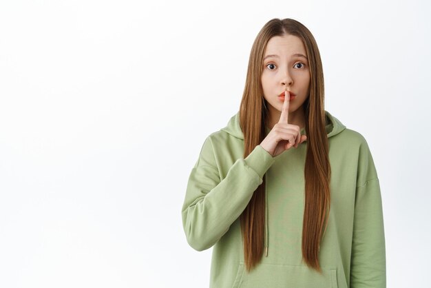 Worried girl asking to stay quiet hold finger on lips and begging to keep secret shushing shhh gesture standing over white background