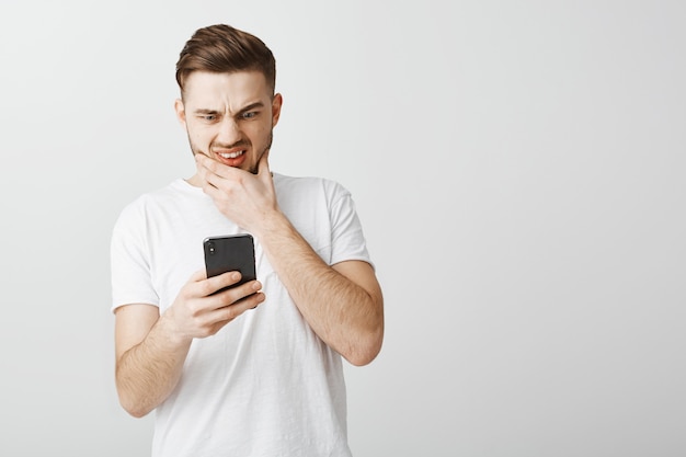Обеспокоенный парень съеживается перед дисплеем смартфона, озабоченно глядя на мобильный телефон
