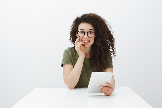 Worried insecure good-looking female student with curly hair, sitting at table with digital tablet