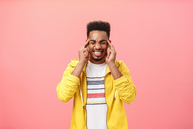 心配している強烈なアフリカ系アメリカ人の男は、こめかみに指を持って目を閉じて歯を食いしばる圧力を処理することができませんピンクの背景に頭痛や片頭痛を感じているストレス下に集中することはできません。