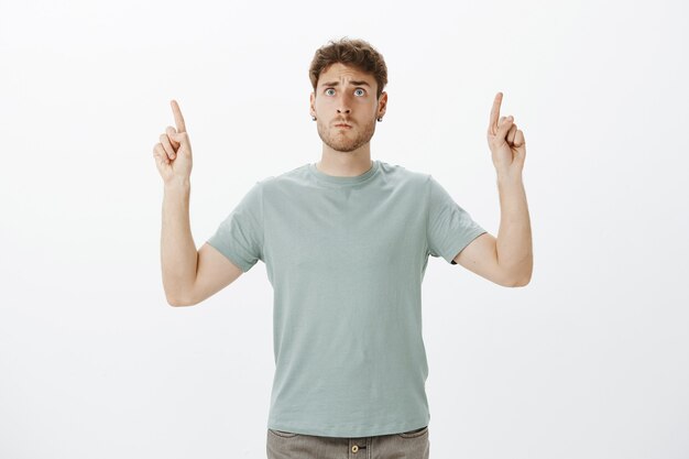 Worried intense funny fair-haired guy in t-shirt, pointing up with raised index fingers and looking at sky with anxious scared expression, worrying for child on playground