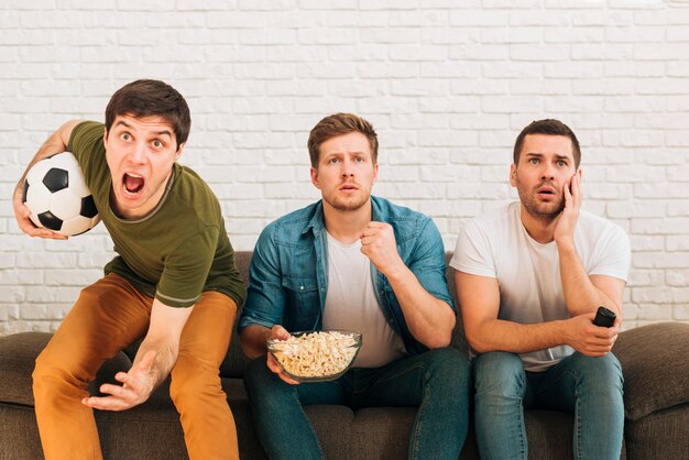 Worried male friends sitting on sofa watching football match on television