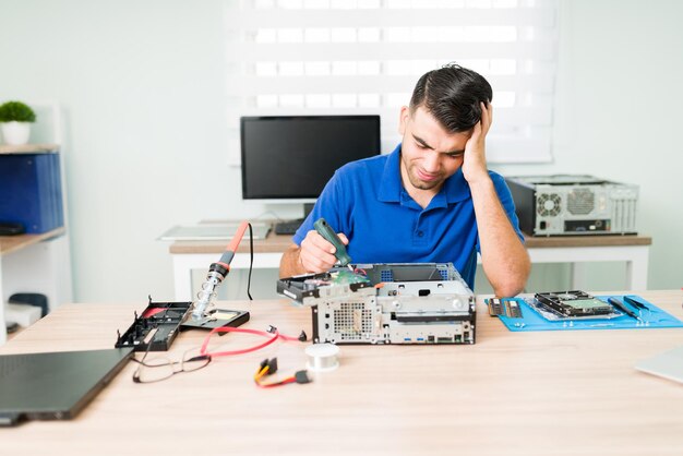 Worried male technician feeling stressed while trying and failing to repair a broken computer at his workshop