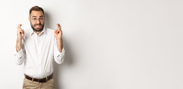 Worried man making a wish cross fingers and hope for relish standing over white background