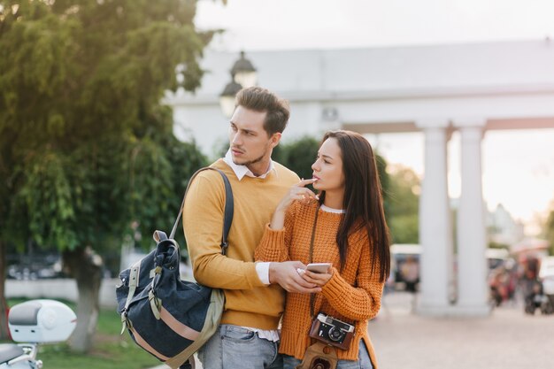 Worried man with backpack looking away, standing beside beautiful woman in orange clothes