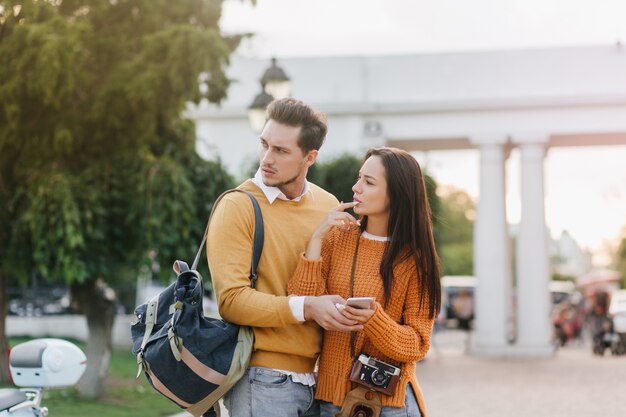 Обеспокоенный мужчина с рюкзаком смотрит в сторону, стоя рядом с красивой женщиной в оранжевой одежде
