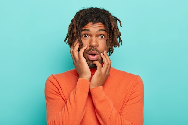 Worried man with dreadlocks, awaits nervously, keeps both palms on cheeks, gasps from fear, wears orange sweater