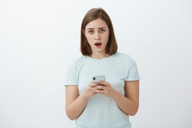 Worried nervous girl asking advice from friend trying text guy she loves standing concerned and troubled over white wall holding smartphone staring unsure and perplexed with opened mouth