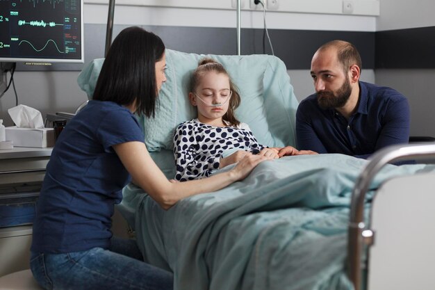 Worried parents sitting beside sick daughter hospitalized in pediatric healthcare facility. Sleeping ill little girl comforted by caring mother and father while resting in patient bed.