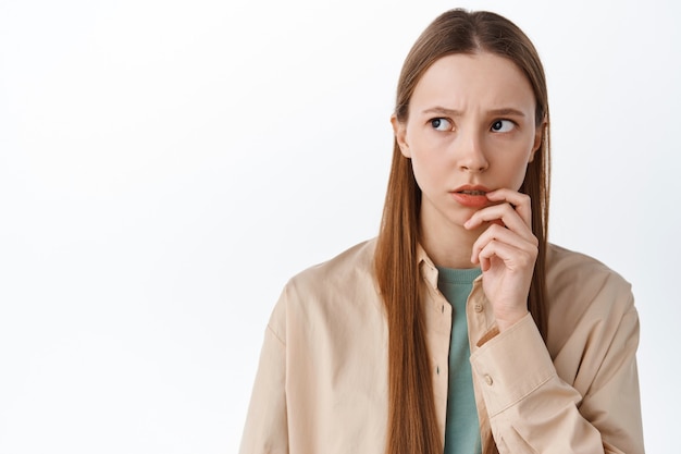 Worried pensive teen student look aside, touching lip nervously and thinking, standing anxious or hesitant against white wall