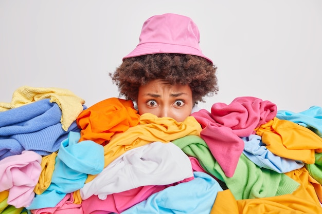 Worried puzzled woman cluttered with unfolded mixed up clothes raises head drom heap of unfolded multicolored laundry 