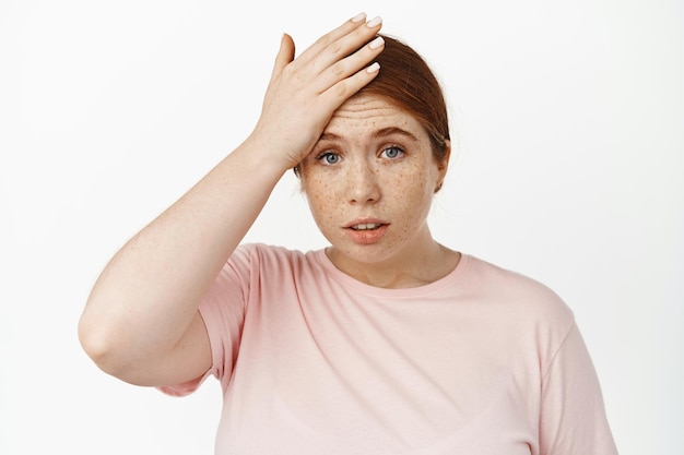 Worried redhead female student slap forehead, looking nervous, forget remember something, slipped her mind, got in trouble, standing in pink t-shirt against white background.