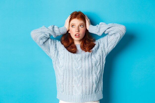 Worried redhead girl standing overwhelmed, holding hands on head in panic and staring left at logo, standing anxious against blue background