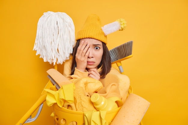 Worried scared Asian housewife looks frightened as notices very dirty room being busy with cleaning surrounded by mops broom brush pile of laundry chemical detergents isolated over yellow wall