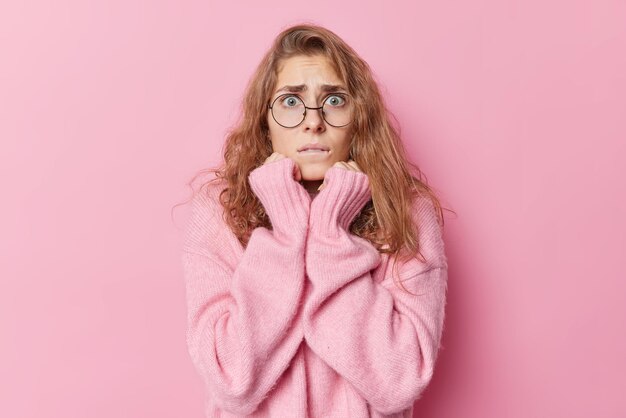 Worried scared young woman looks anxious afraids of something scarying bites lips wears big round spectacles and warm cashmere jumper isolated over pink background smells problems looks intense