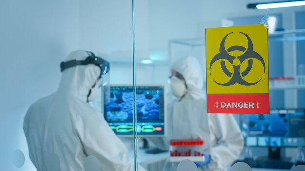 実験室の危険な領域で働いているガラスの壁の後ろで話しているppeスーツの心配している科学者