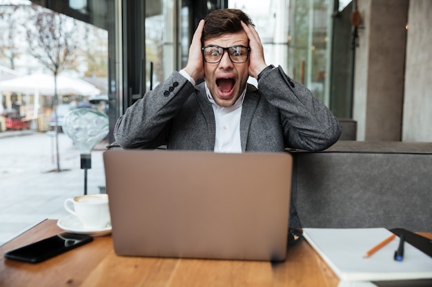 Worried screaming businessman in eyeglasses sitting by the table in cafe and holding head