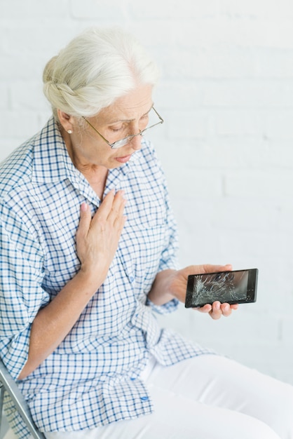 Worried senior woman looking at smartphone with broken screen against white wall