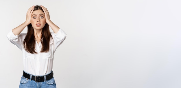 Free photo worried and shocked young woman looking stunned at disaster holding hands on head panicking standing