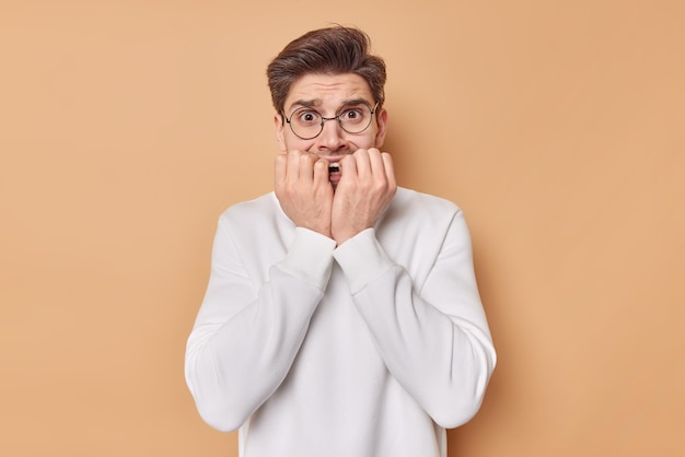 Free photo worried stressed man bites finger nails afraids of something feels embarrassed while akes decision expresses negative emotions wears white jumper isolated over brown background. human reactions