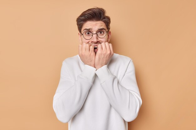 Worried stressed man bites finger nails afraids of something feels embarrassed while akes decision expresses negative emotions wears white jumper isolated over brown background. Human reactions