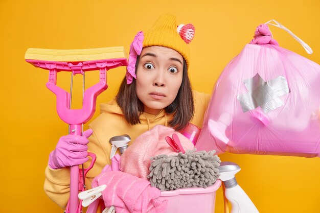 Worried surprised Asian woman helps parents to do housework collects garbage in polythene bag washes floor with mop uses different cleaning tools and detergents isolated over yellow studio wall