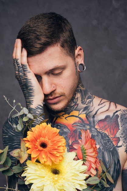 Worried tattooed young man with nose and ear piercing holding flower in hand