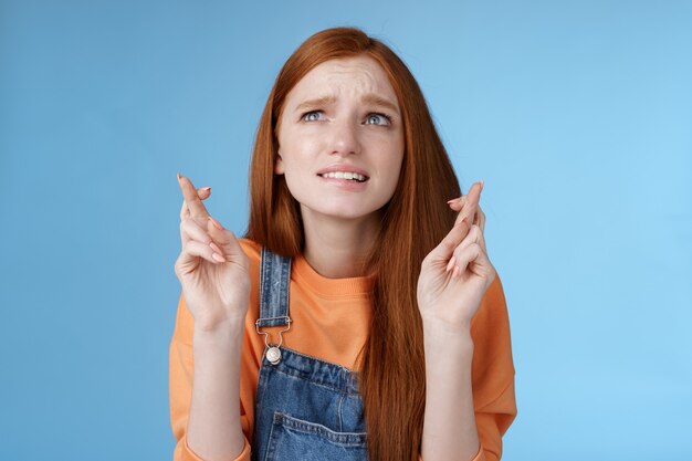 Worried unconfident young redhead intense cute girl frowning nervously waiting improtant moment cross fingers good luck bite lip anxiously frowning look upper left corner praying wish come true