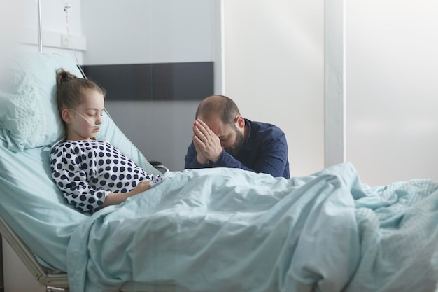 걱정스러운 젊은 아버지는 병실에 있는 동안 입원한 아픈 환자를 위해 기도합니다. 산소 튜브를 통해 호흡하면서 병에서 회복하는 아픈 어린 소녀 딸.