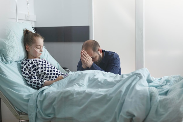 Worried uneasy young father praying for hospitalized sick patient girl while in medical room. Ill little girl daughter recovering from sickness while breathing through oxygen tube.