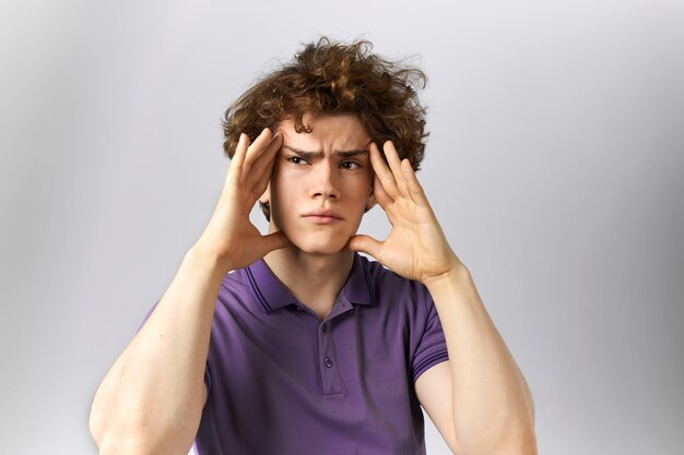 Worried upset young man with curly hair having exhausted tired look massaging temples trying to soothe pain while suffering from migraine or headache. Sad guy being depressed because of problems