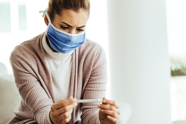 Worried woman wearing protective face mask and measuring her temperature at home