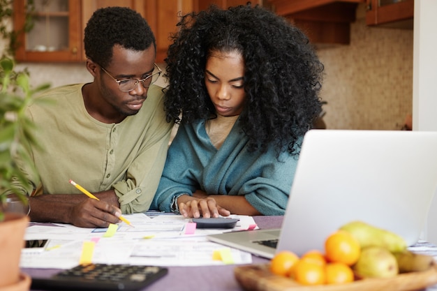 Worried young African family of two facing financial difficulties. Unhappy woman with Afro hairstyle using calculator while doing paperwork with her husband who is filling in papers with pencil