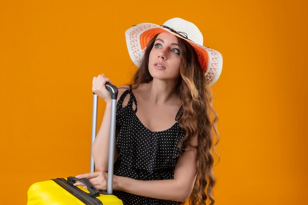 Worried young beautiful girl in dress in polka dot in summer hat standing with suitcase looking aside over yellow background