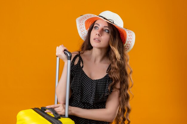 Worried young beautiful girl in dress in polka dot in summer hat standing with suitcase looking aside over yellow background