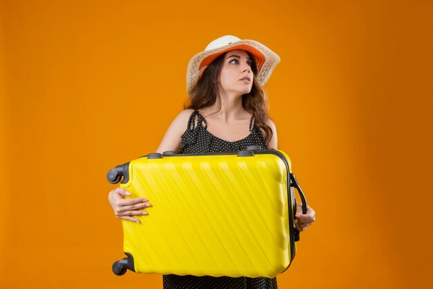 Worried young beautiful traveler girl in dress in polka dot in summer hat holding suitcase looking aside standing over yellow background