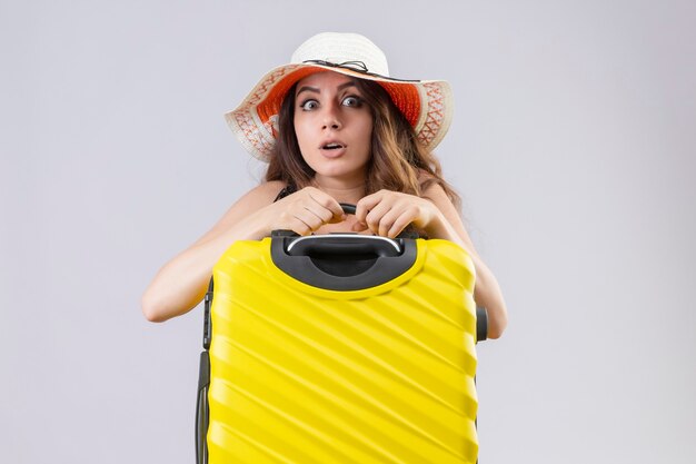Worried young beautiful traveler girl in dress in polka dot in summer hat holding suitcase looking at camera standing over white background