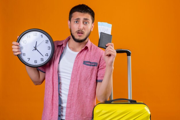 Взволнованный молодой красивый путешественник, держащий авиабилеты и часы, смотрит в камеру с растерянным выражением лица, стоя с чемоданом на оранжевом фоне